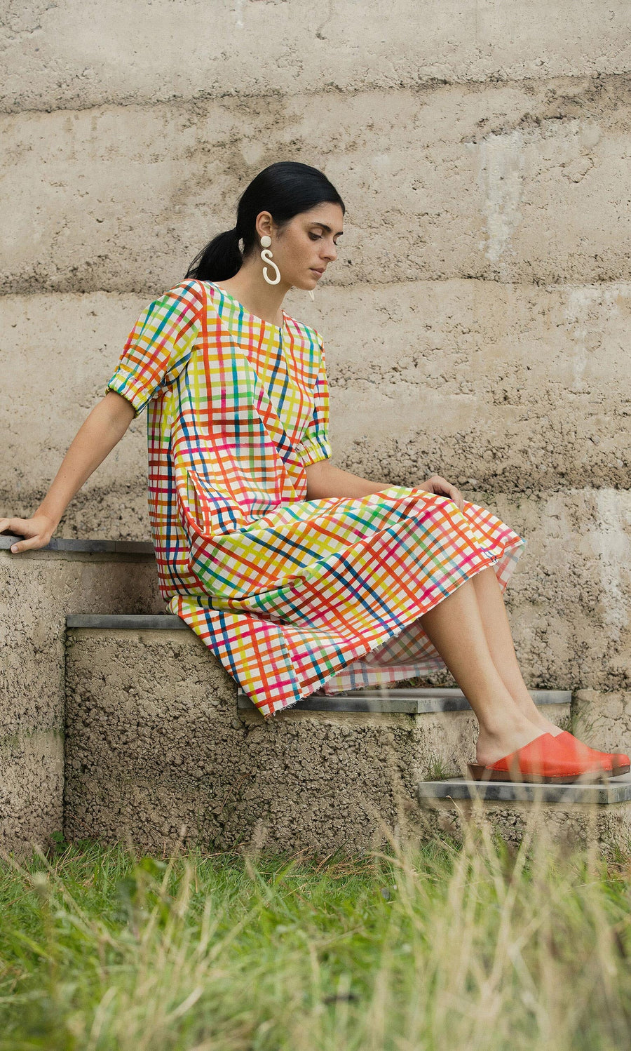 Plümo- Woman in Thea Dress, a hand-dyed check cotton-poplin, sits on steps with statement earrings and orange shoes.