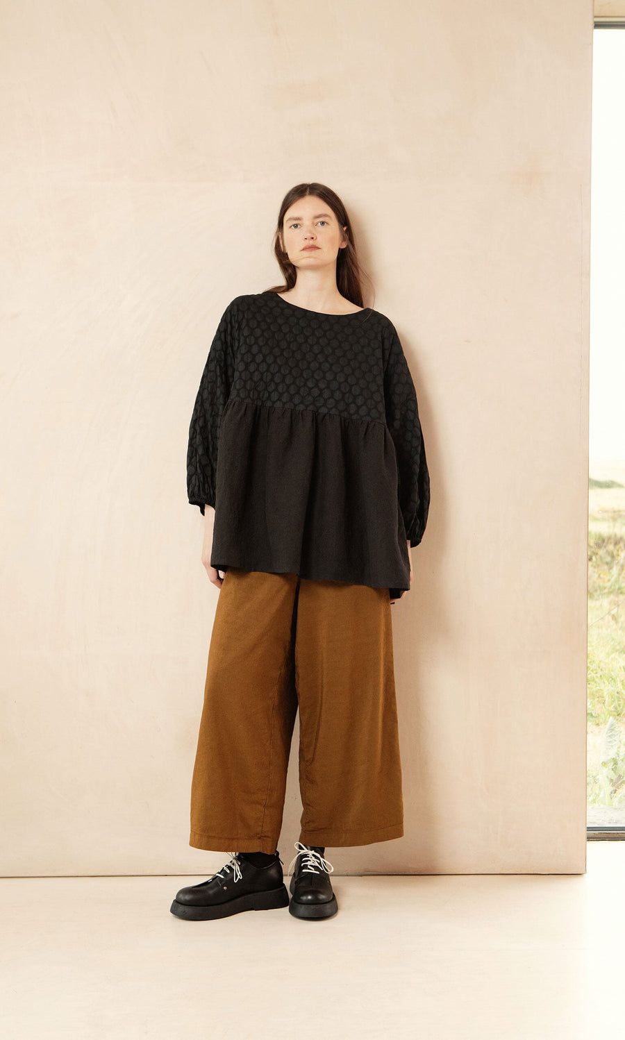 Plümo- Woman in the Tuppence Blouse and wide brown pants stands against a beige wall, wearing black shoes.