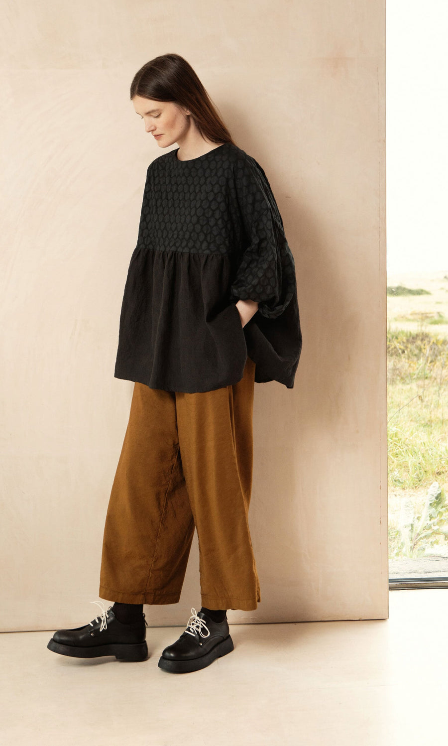 Plümo- A woman stands indoors in the Tuppence Blouse with wide brown pants and black shoes, looking down and smiling.