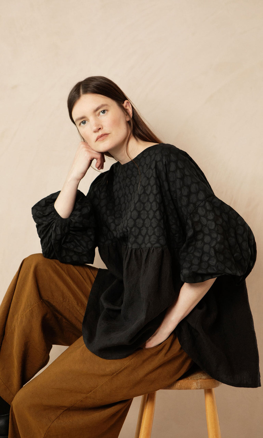 Plümo- Woman with long brown hair sits on a stool, wearing the Tuppence Blouse and brown pants, head resting on her hand.