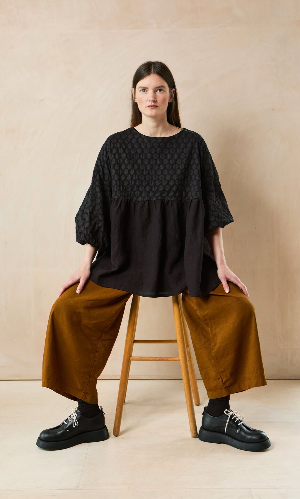 Plümo- Woman with long brown hair sits on a stool, wearing the Tuppence Blouse and wide mustard pants with black shoes.