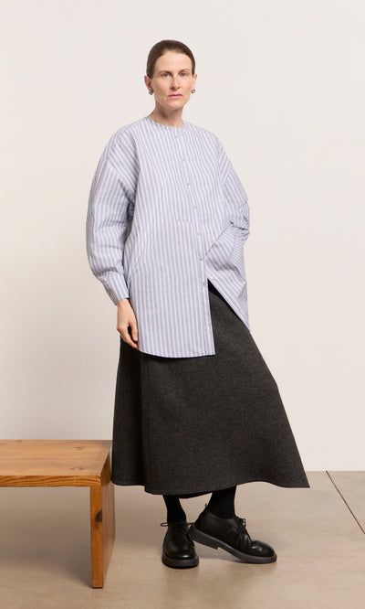 Plümo  Person in a chambray blue Tuxedo Shirt and dark skirt stands by a wooden bench, looking at the camera.