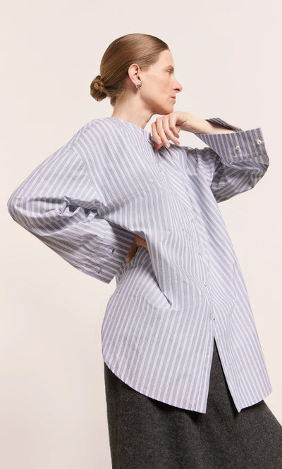 Plümo  Woman in a blue striped Tuxedo Shirt and dark skirt, posing sideways with hair styled in a low bun.