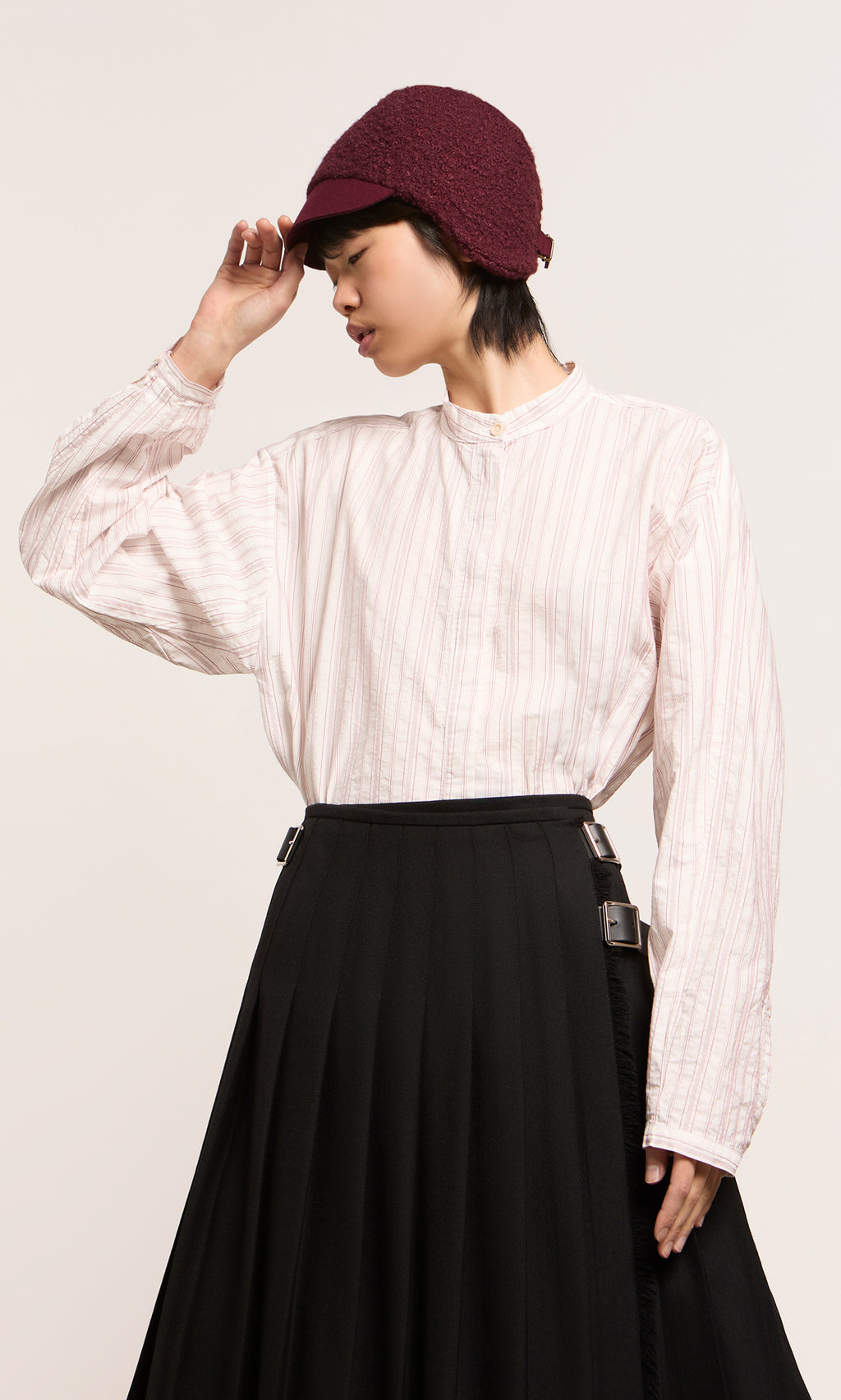 Plümo  Model wears loose white striped shirt, black skirt, and maroon Velo Wool Cap with ear flaps against a plain background.