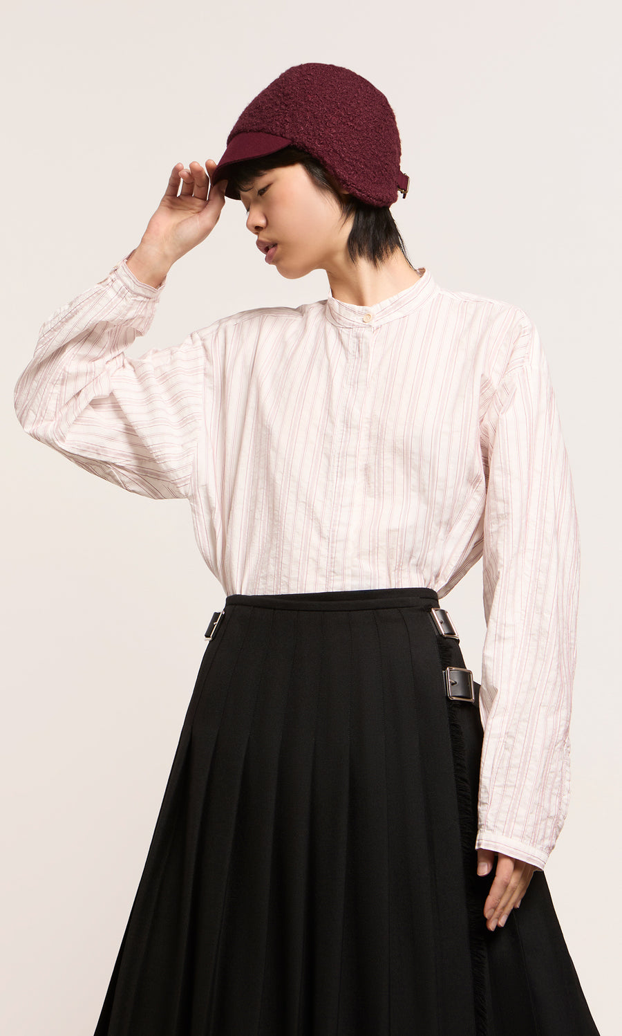 Plümo  Model wears loose white striped shirt, black skirt, and maroon Velo Wool Cap with ear flaps against a plain background.