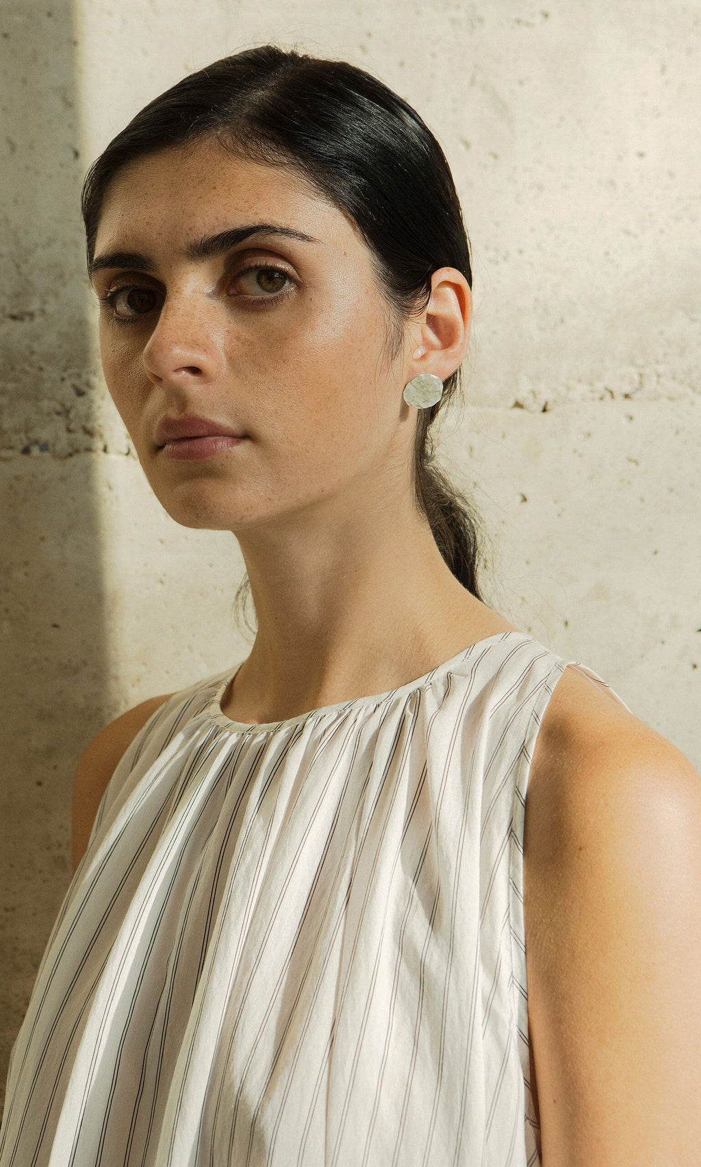 Plümo- Woman with dark hair in a low ponytail, wearing Noreen Studs and a white striped top, stands by a stone wall.