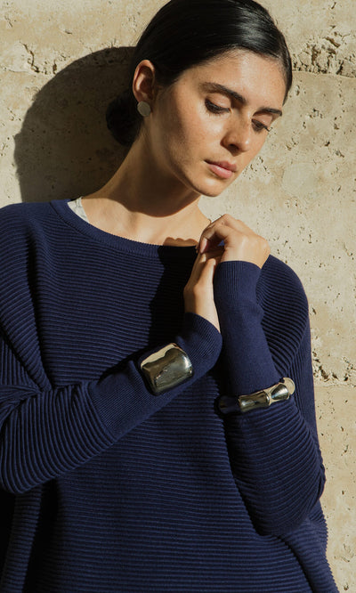 Plümo- Woman in a navy ribbed sweater wearing the Tunner Cuff, standing against a textured beige wall in sunlight.