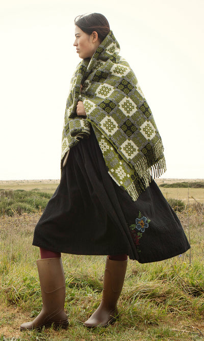 Plümo- Woman in boots draped in an Apple Green Welsh Blanket stands in a grassy field on a cloudy day.