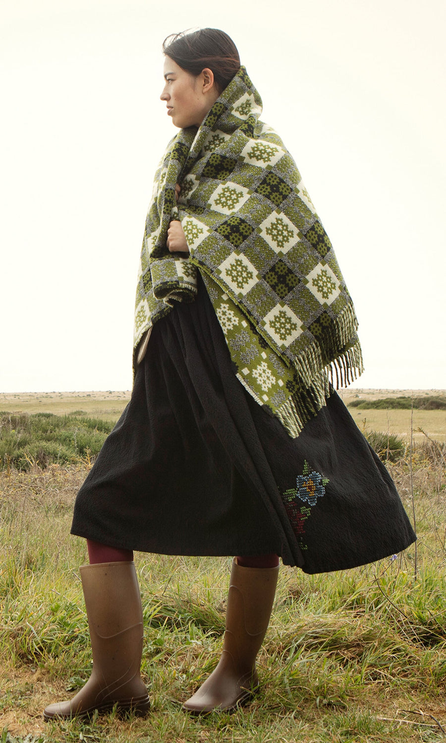 Plümo- Woman in boots draped in an Apple Green Welsh Blanket stands in a grassy field on a cloudy day.