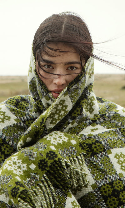 Plümo- A woman stands outdoors, wind in her hair, wrapped in a Welsh Blanket - Apple Green.