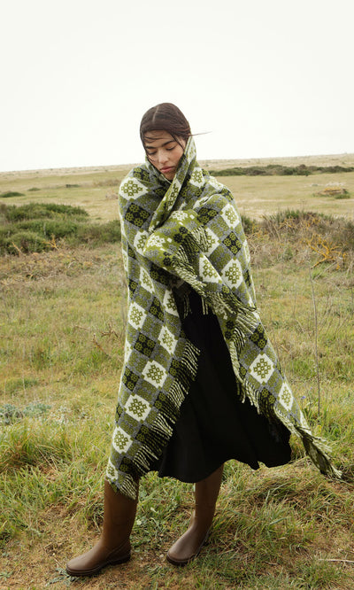 Plümo- A woman stands in a grassy field, eyes closed, wrapped in an Apple Green Welsh Blanket, wearing boots and dark clothes.