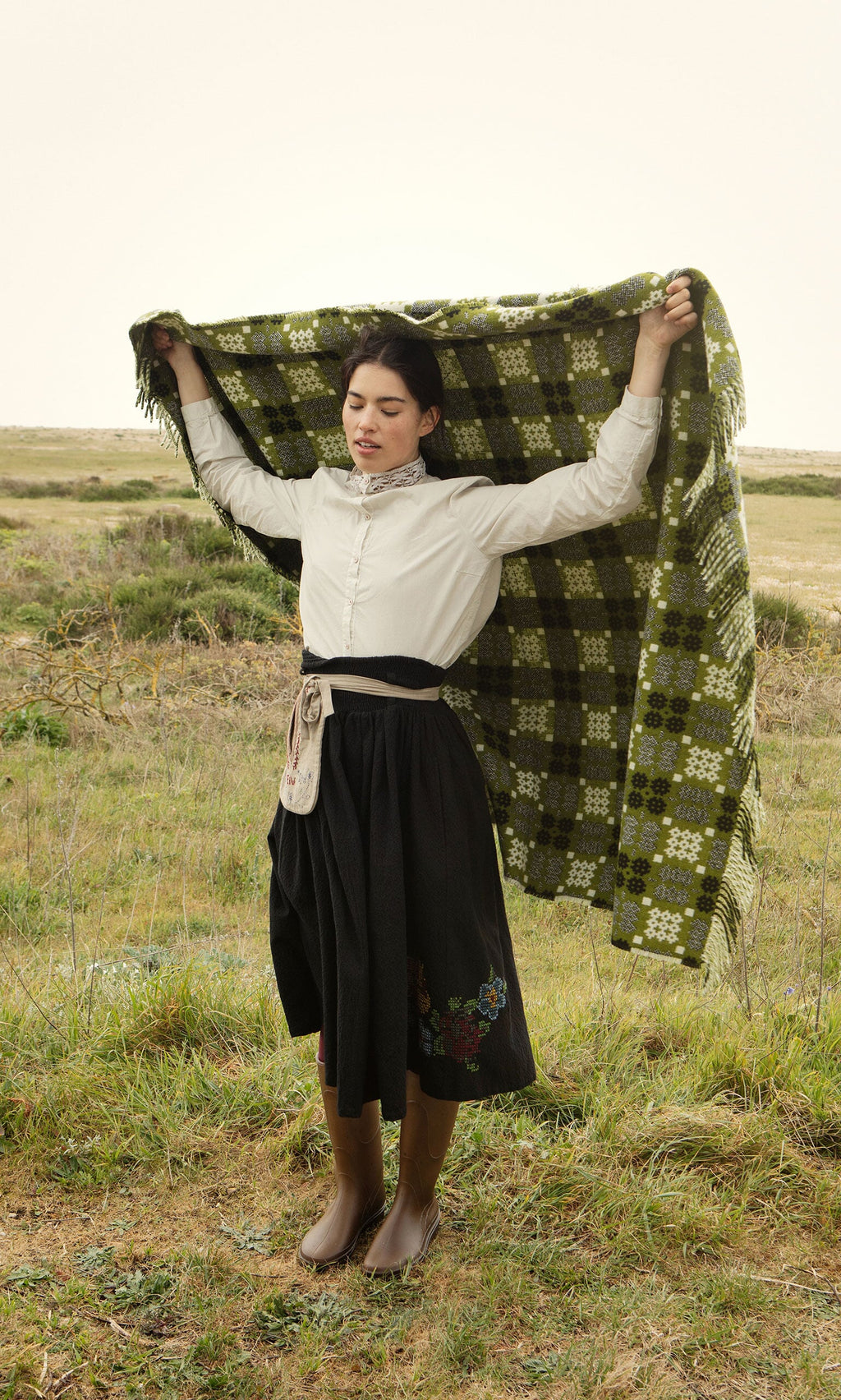 Plümo- A woman in a field holds the Welsh Blanket - Apple Green, wearing a white blouse and black skirt.