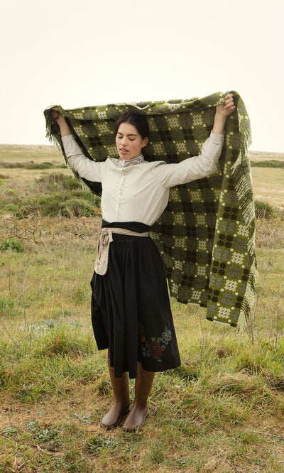 Plümo- A woman in a field holds the Welsh Blanket - Apple Green, wearing a white blouse and black skirt.