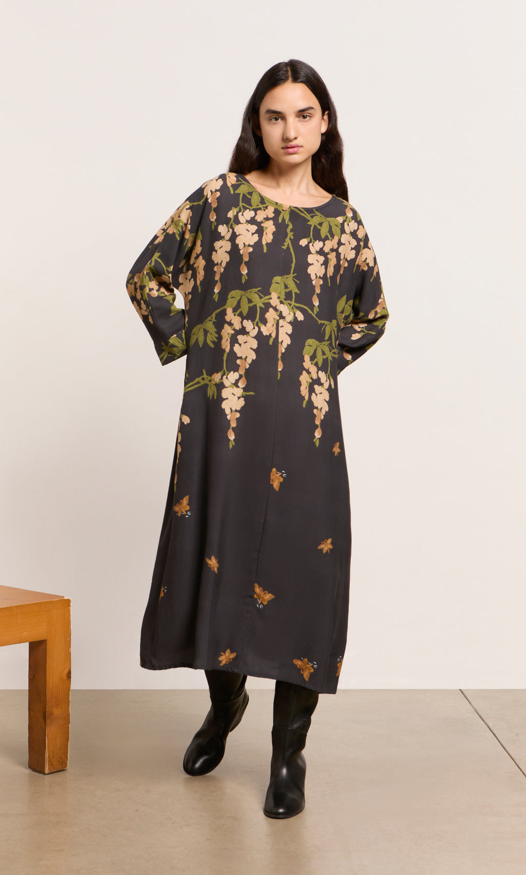 Plümo  Woman indoors in a green and tan Whisteria Dress with black boots, featuring botanical-inspired patterns.