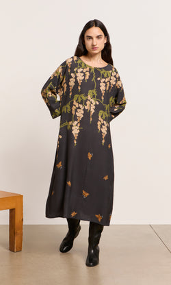 Plümo  Woman indoors in a green and tan Whisteria Dress with black boots, featuring botanical-inspired patterns.