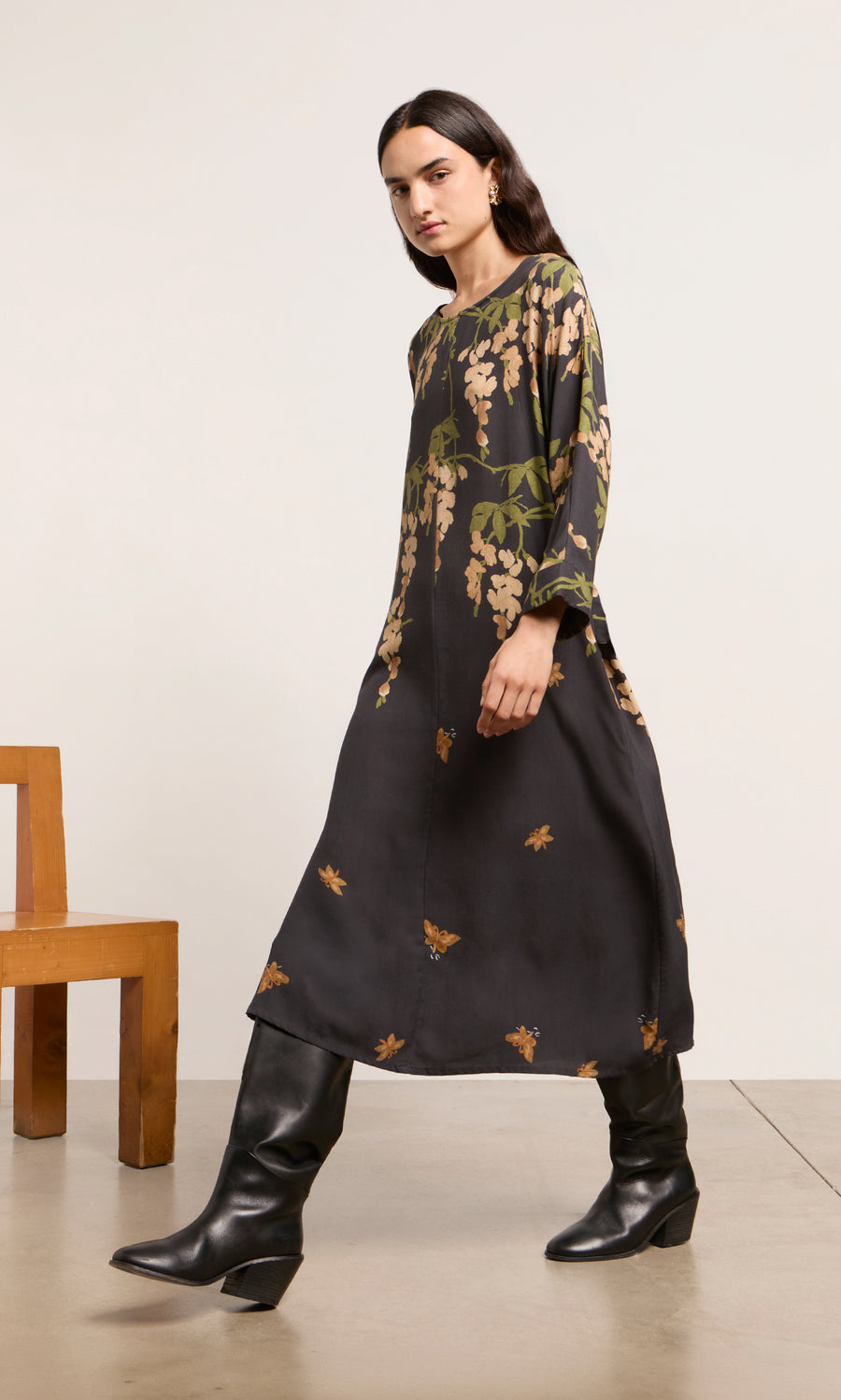 Plümo  Woman in a Whisteria Dress and tall black boots walking indoors near a wooden chair.