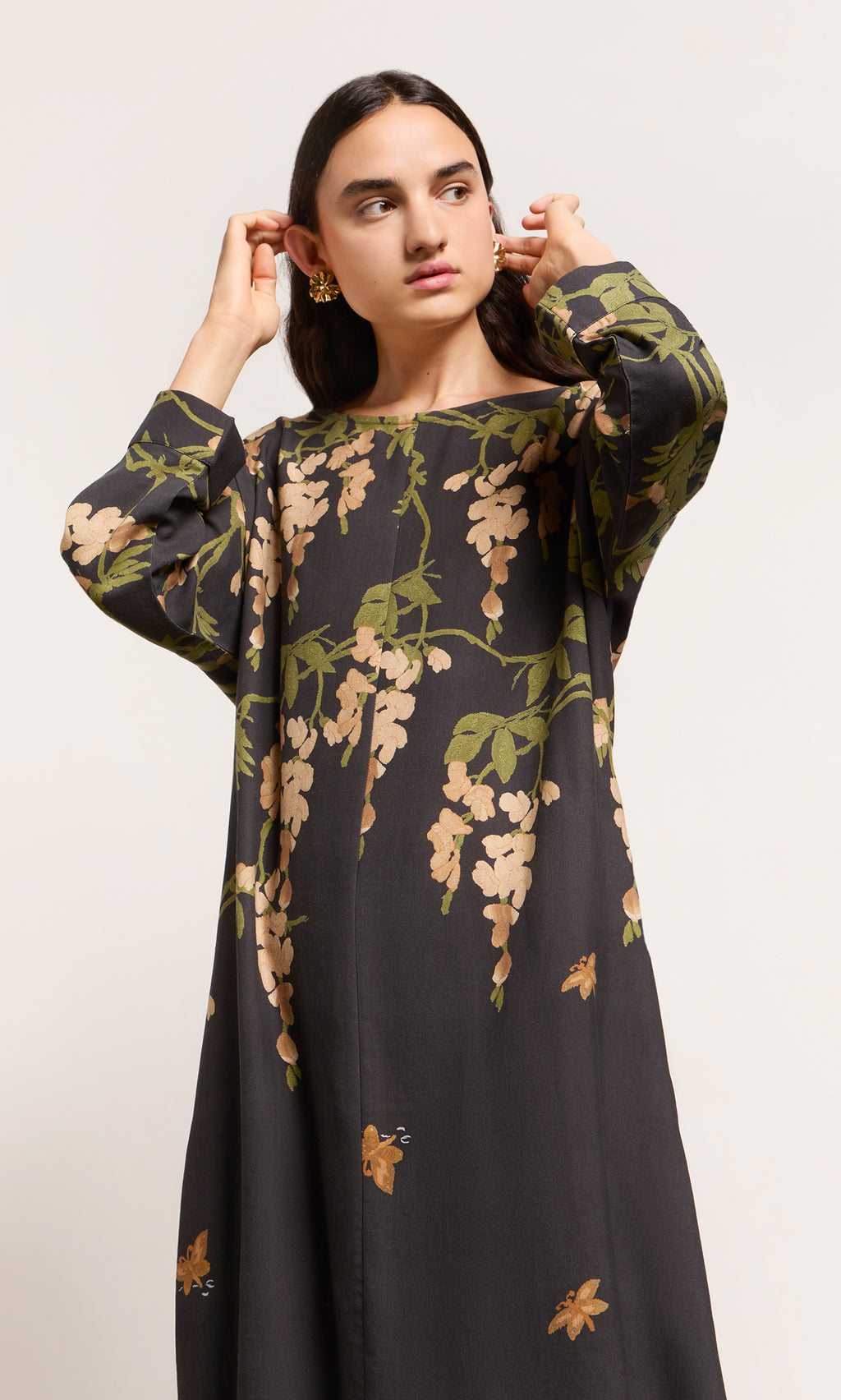 Plümo  Woman in a Whisteria Dress adjusts her earrings, gazing off to the side against a simple background.