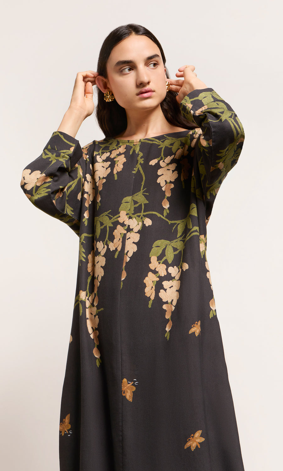 Plümo  Woman in a Whisteria Dress adjusts her earrings, gazing off to the side against a simple background.