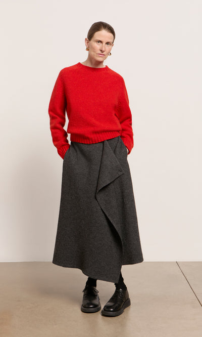 Plümo  Person in a red Wicklow Crewneck sweater, gray wrap skirt, black tights, and shoes stands against a plain background.