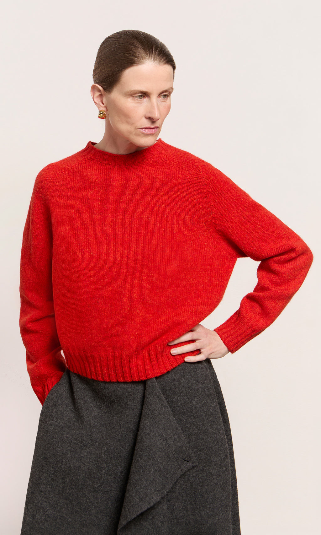 Plümo  Woman wears the Wicklow Crewneck - Red Fleck sweater and a gray skirt, standing with one hand on her hip, looking aside.