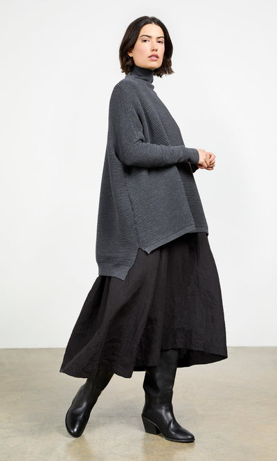 Plümo- Woman wears the Jack Rollneck - Graphite with a black skirt and boots, standing against a plain background.