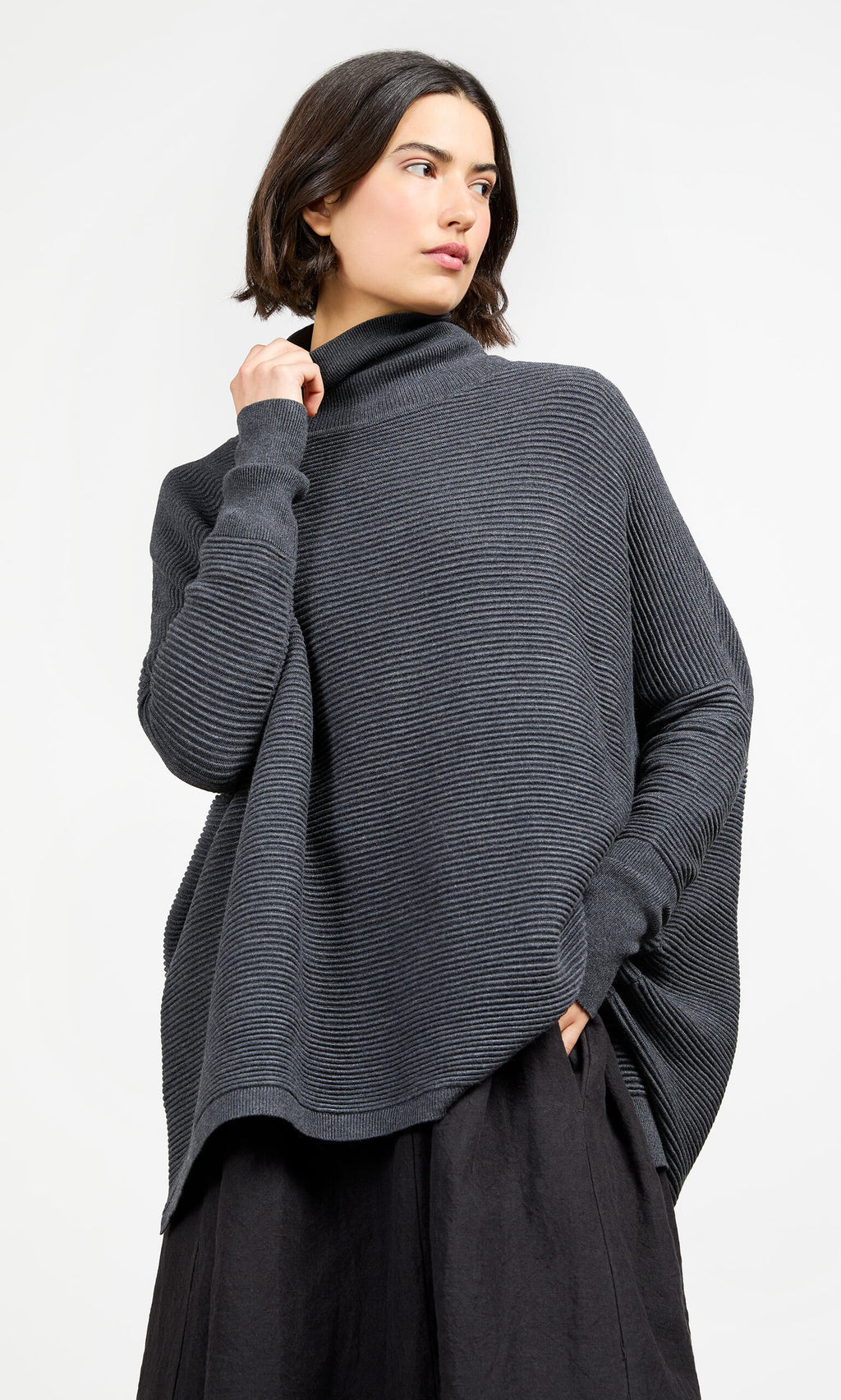 Plümo- Woman in Jack Rollneck - Graphite, an oversized ribbed sweater, and black skirt, looking to the side.