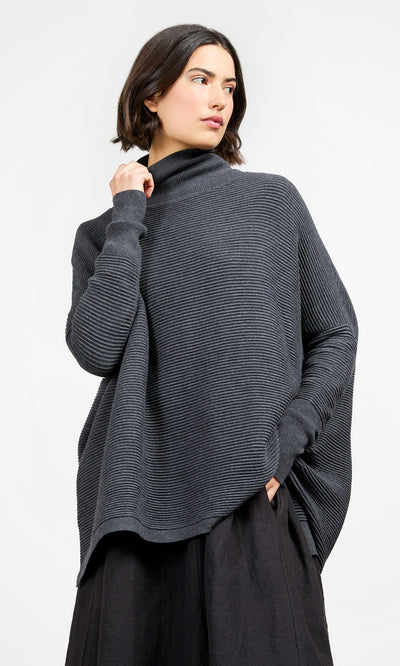Plümo- Woman in Jack Rollneck - Graphite, an oversized ribbed sweater, and black skirt, looking to the side.