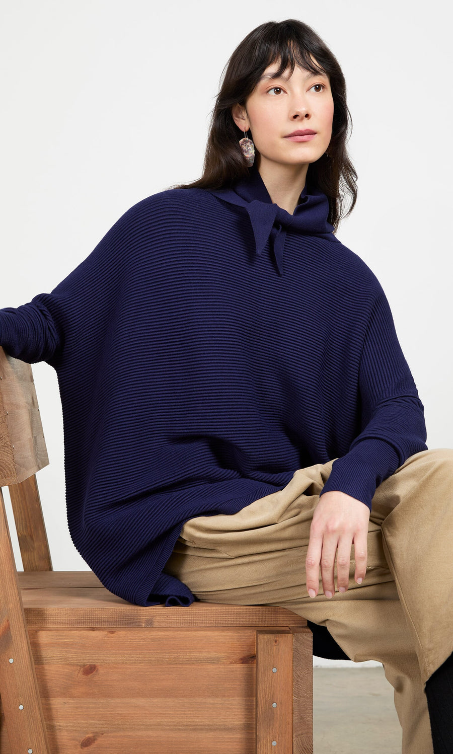 Plümo- Woman in a navy Jona Necktie and tan pants sits calmly on a wooden chair, looking to the side.