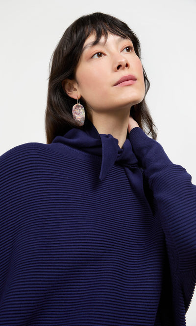 Plümo- Woman with long dark hair in a ribbed navy Jona Necktie top and marbled oval earring, looking upward.