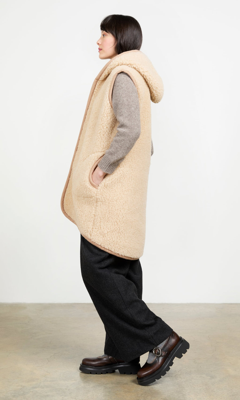 Plümo- Woman in the Long Rambler Gilet - Natural, gray sweater, black pants, and brown shoes stands sideways by a white wall.