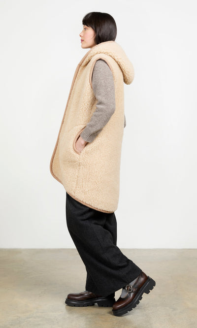 Plümo- Woman in the Long Rambler Gilet - Natural, gray sweater, black pants, and brown shoes stands sideways by a white wall.