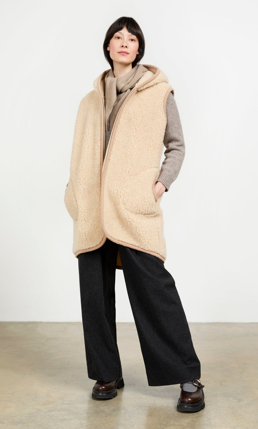 Plümo- Someone wearing the Long Rambler Gilet - Natural, a gray sweater, wide-leg pants, and brown shoes stands against a plain backdrop.