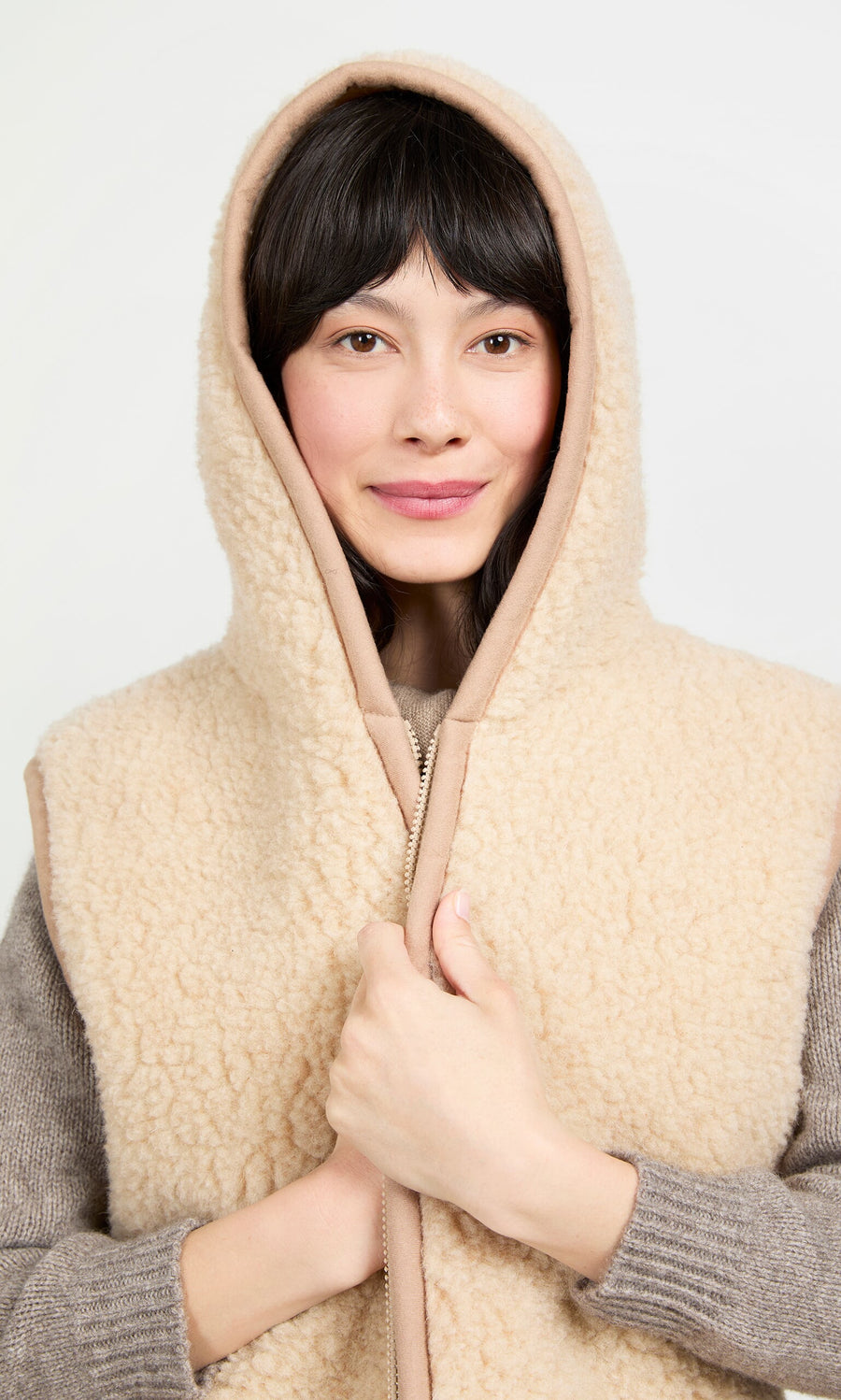 Plümo- Woman with straight black hair smiles, wearing the Long Rambler Gilet - Natural over a gray sweater.