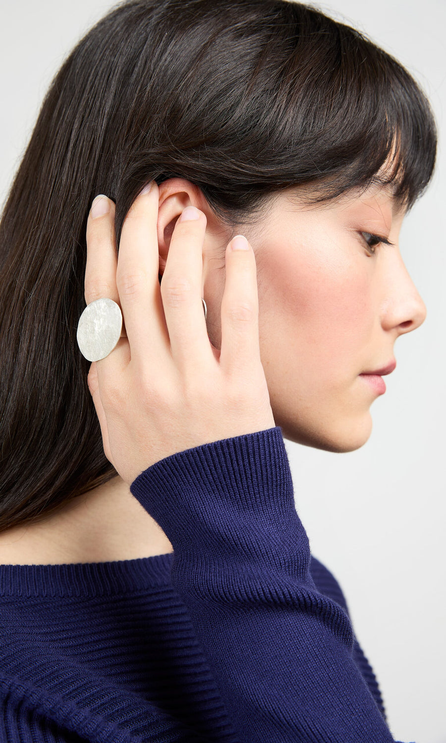 Plümo- A woman in a navy sweater wears our handmade Eco Silver Pebble Ring, touching her ear.