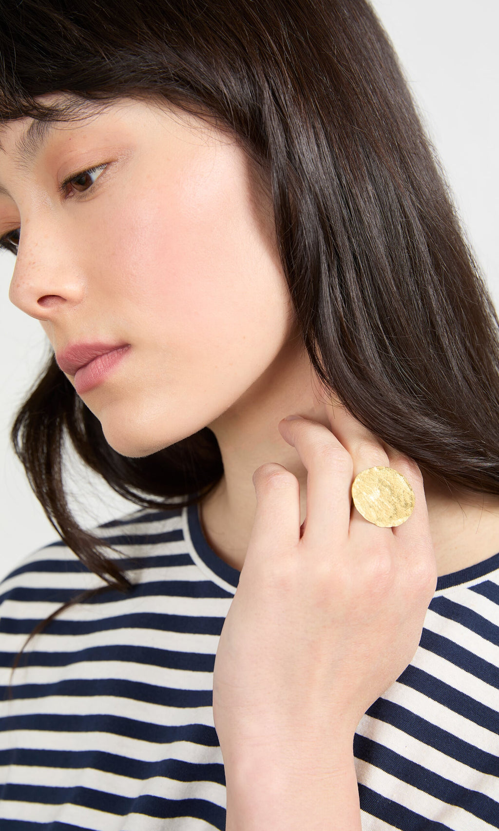 Plümo- Woman with long dark hair in a striped shirt wears the Brass Pebble Ring, gazing down thoughtfully.