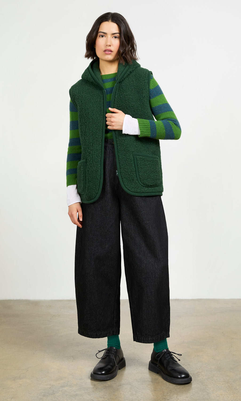 Plümo- Person wears a green striped sweater, Wool Gilet - Moss Green, wide-leg black pants, and black shoes against a plain background.