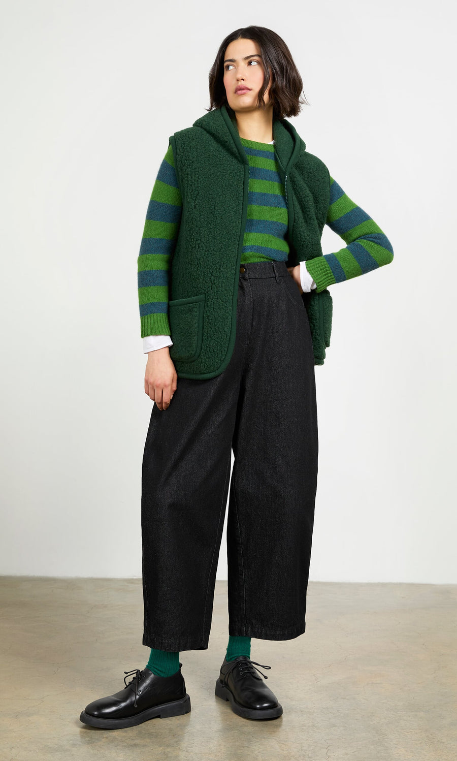 Plümo- Woman in a moss green Wool Gilet over striped sweater, black wide-leg pants, green socks, and black shoes indoors.