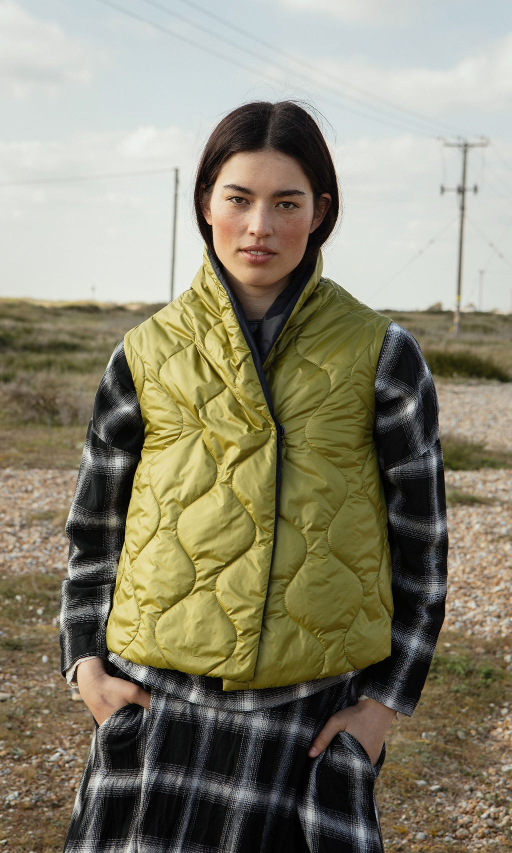Plümo- Woman outdoors wears the Acacia Gilet, a lightweight green quilted vest, over a black-and-white plaid dress.
