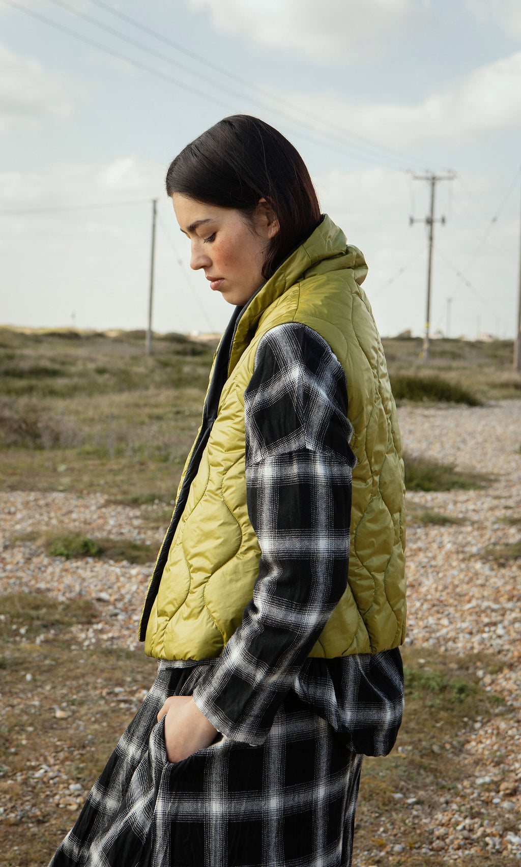 Plümo- A woman in an Acacia Gilet and plaid dress stands outdoors on rocky terrain, looking down thoughtfully.