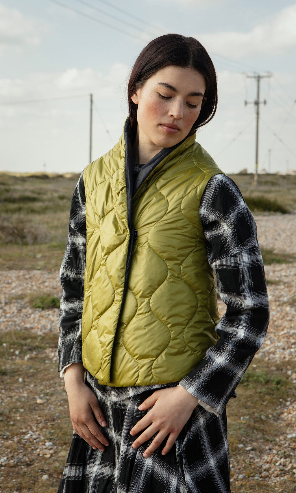 Plümo- Woman in the Acacia Gilet and a plaid dress stands outdoors on a gravel path beneath power lines.
