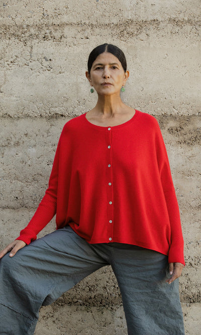 Plümo- A woman confidently poses by a beige wall in the Alma Cardigan - Red, featuring pearly buttons.
