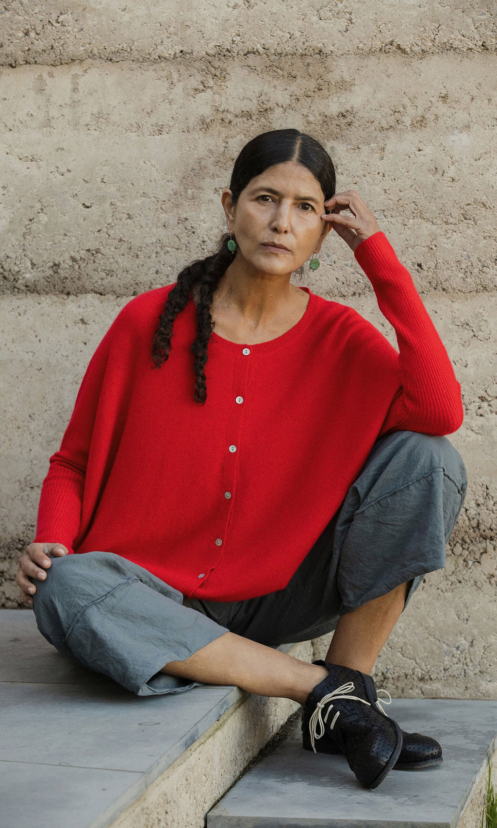 Plümo- Woman in the Alma Cardigan - Red sits on steps, looking thoughtfully at the camera.
