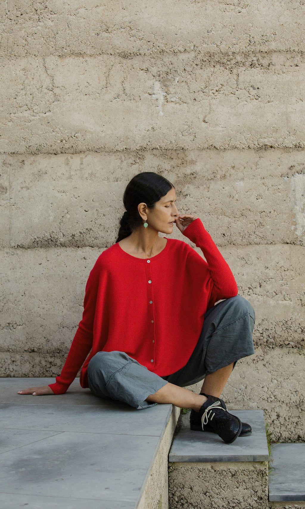 Plümo- A woman in the Alma Cardigan - Red sits on a concrete ledge, gazing to the side.