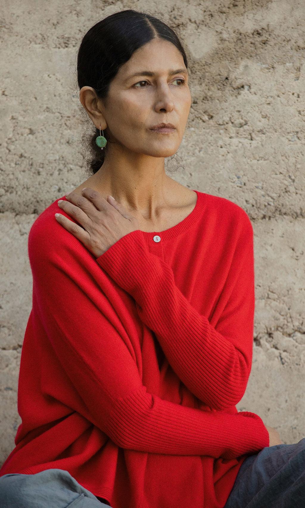 Plümo- A woman with dark hair in the Alma Cardigan - Red sits by a textured wall, gazing thoughtfully to the side.