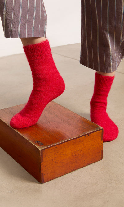 Plümo  Wearing Alpaca Socks - Postbox Red, a person in brown pinstripe pants stands on a wooden block.
