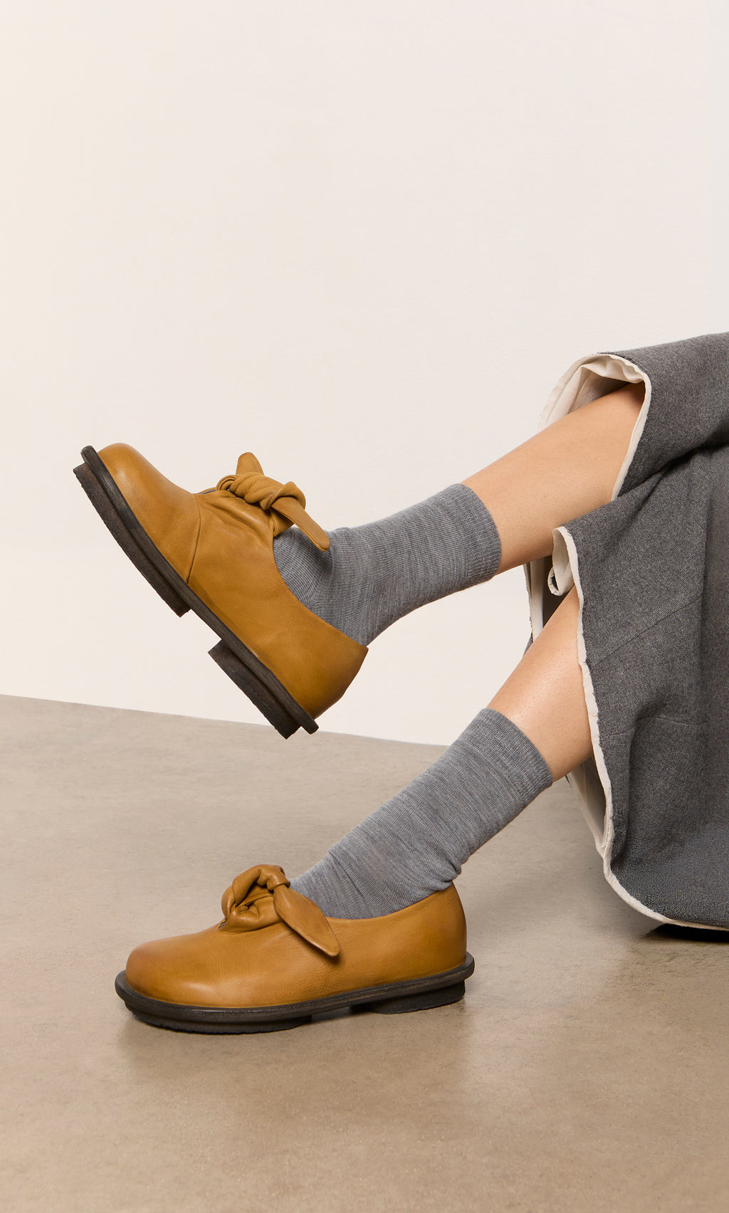 Plümo  Person wearing Alpaca Socks - Slate Grey, brown bow leather shoes, gray skirt, sitting on a tan floor.