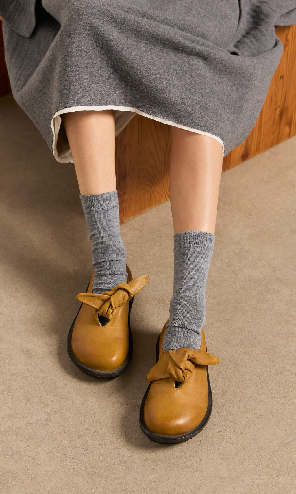 Plümo  Person in gray skirt and socks wears Amber Leather Shoes while sitting on a wooden bench.