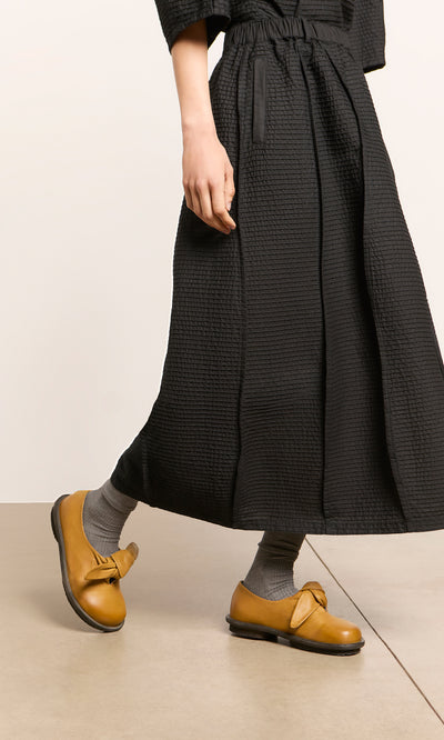 Plümo  Individual wearing a long black skirt, gray socks, and Amber Leather Shoes walks indoors.