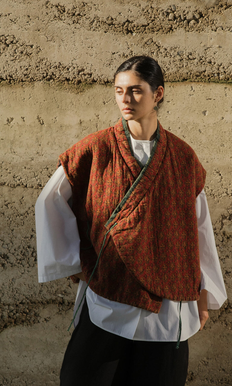 Plümo- Person in the Ansel Wrap Jacket and white shirt stands by a sunlit beige wall, looking to the side.