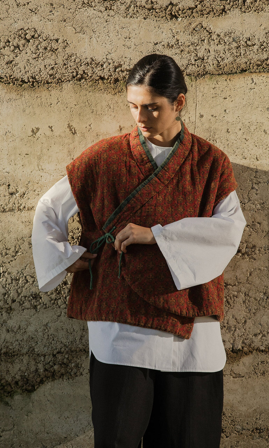 Plümo- Person in a red, block-printed Ansel Wrap Jacket ties the sash against a textured earth-toned wall.